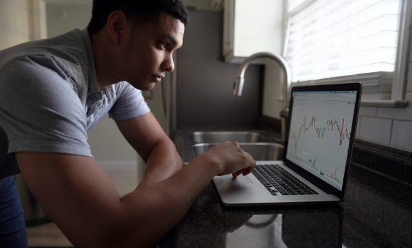 A man stands at his kitchen counter studying investments on his laptop