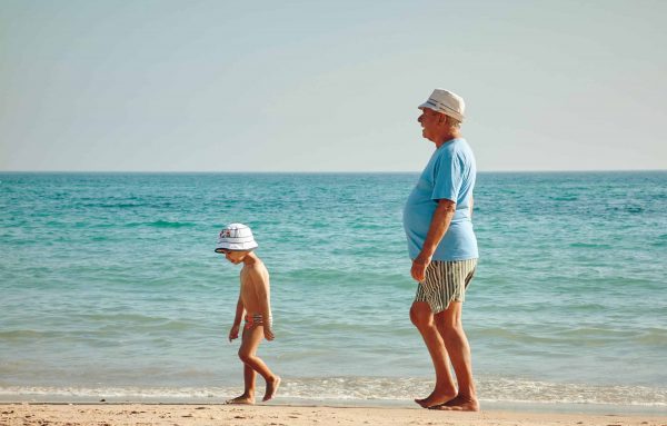 a young child walks on the beach followed by an elderly man in an article about the need to start saving for retirement at a young age