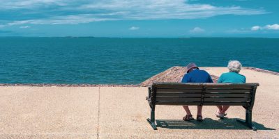 Elderly couple sit on a park bench looking out on a bright blue ocean in a story about income in retirement