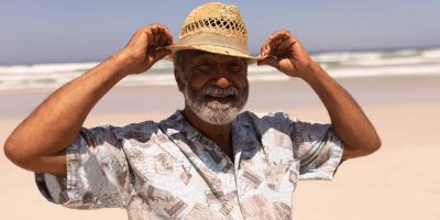 An older man adjusts his hat on a beach, looking retired