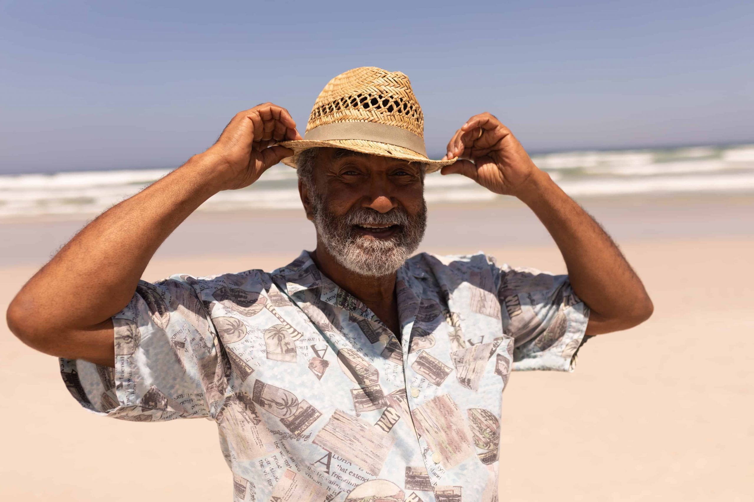 An older man adjusts his hat on a beach, looking retired