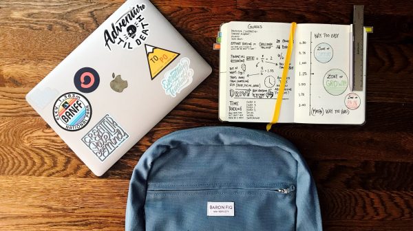 A birds eye view looking down on a laptop covered in stickers, a notebook and a backpack, symbolizing academics and the college application process