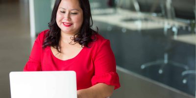 A professional woman in a red dress works at a laptop while smiling, looking empowered